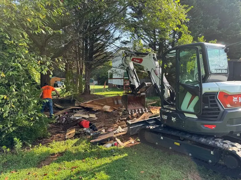 Bobcat E55 excavator with a Land Pros Land Management sticker breaking down a wooden structure in a backyard demolition project